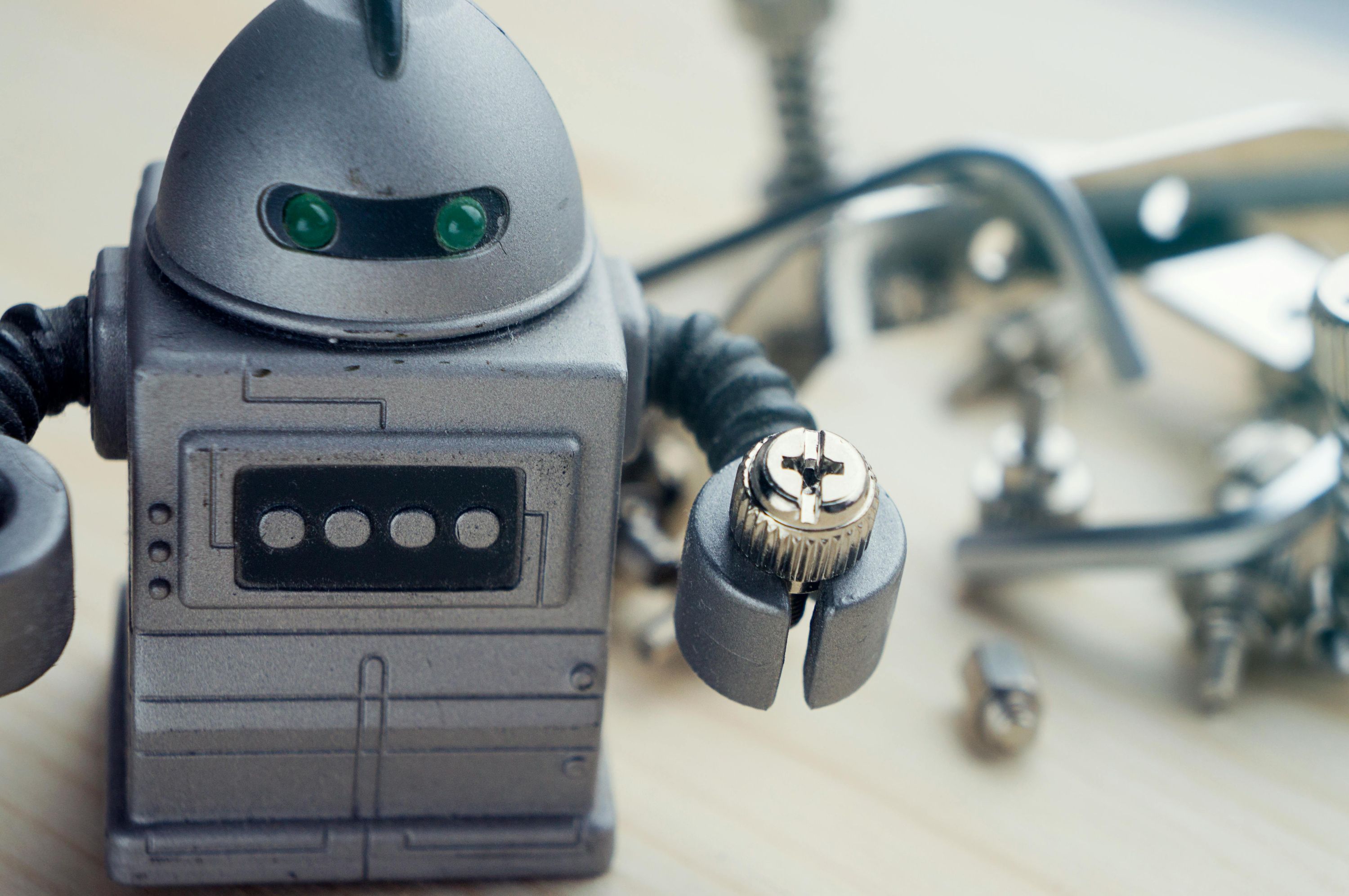 Close-up of a silver toy robot holding a small screw in its claw, with other mechanics blurred in the background.