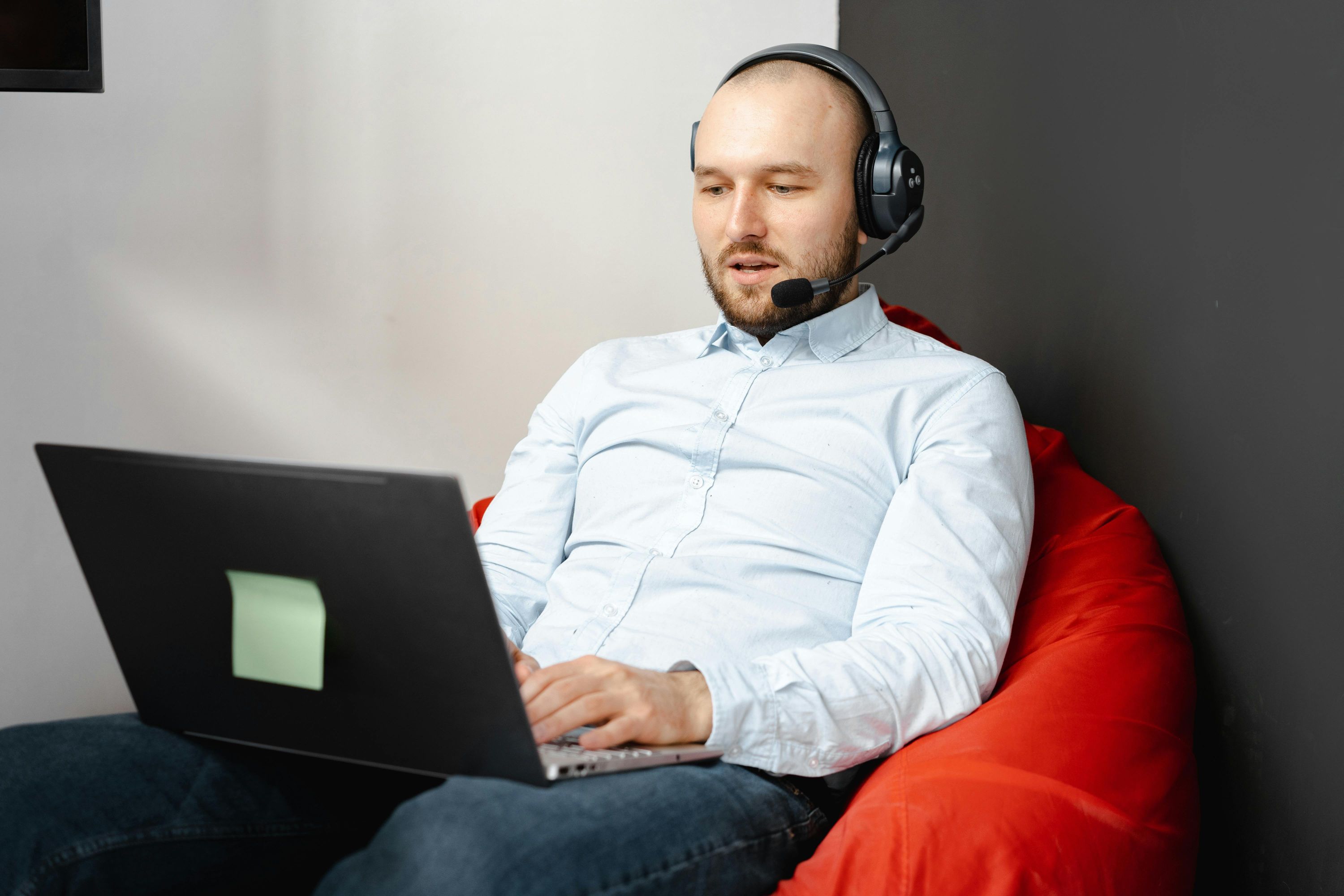 A man wearing a light blue shirt and a headset with a microphone sits on a red beanbag chair, typing on a laptop.