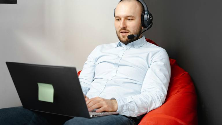 Remote Marketers South Africa: Man in Headset Working on Laptop A man wearing a light blue shirt and a headset with a microphone sits on a red beanbag chair, typing on a laptop.