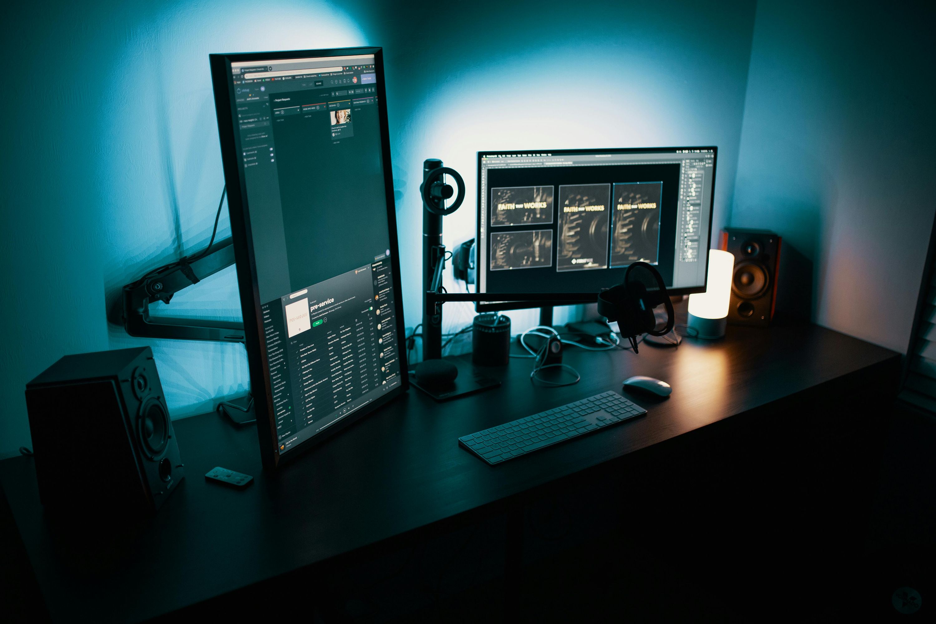 Modern remote work desk setup with two monitors showing project management software and design files, keyboard, mouse, and speakers under blue lighting.