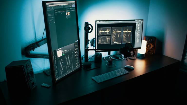 Remote Team Monitoring with Dual Monitor Setup Modern remote work desk setup with two monitors showing project management software and design files, keyboard, mouse, and speakers under blue lighting.