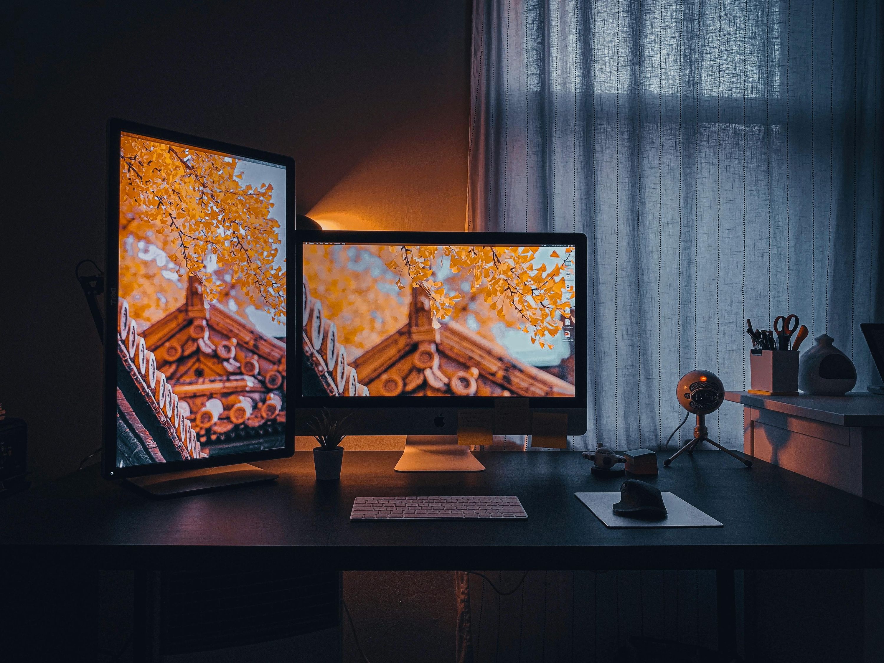 Well-lit dual monitor setup on a desk with a keyboard, mouse, small plant, and microphone, next to a window with striped curtains.