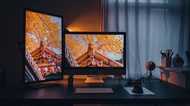 Remote Workstation Setup for Admin Talent Well-lit dual monitor setup on a desk with a keyboard, mouse, small plant, and microphone, next to a window with striped curtains.