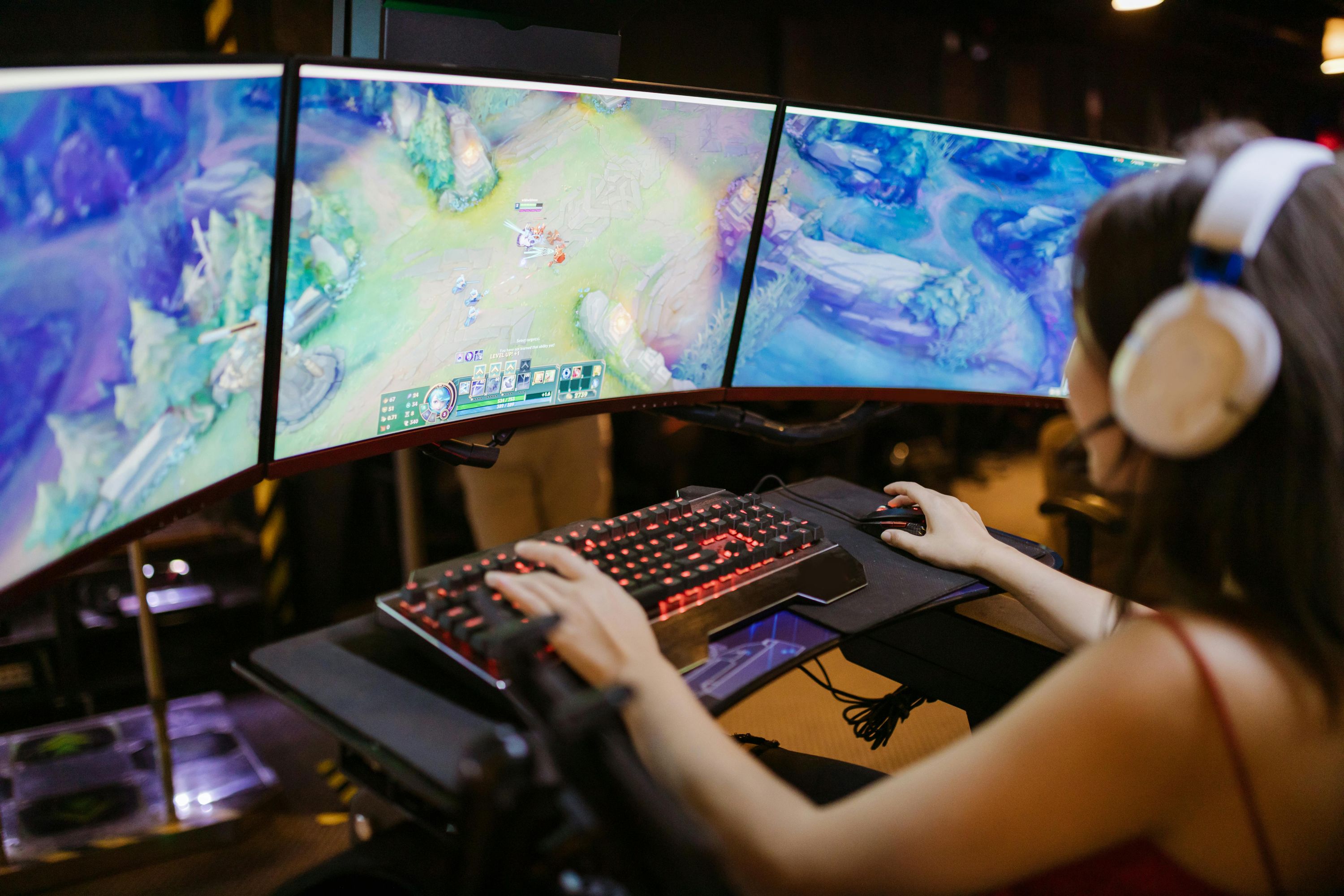 Top-down view of a person wearing headphones, actively gaming on a multi-monitor setup with a glowing keyboard and mouse, immersed in a fantasy video game.