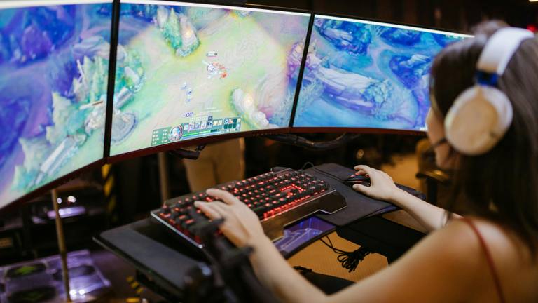 Marketing Consultant for Scaling Business Success Top-down view of a person wearing headphones, actively gaming on a multi-monitor setup with a glowing keyboard and mouse, immersed in a fantasy video game.
