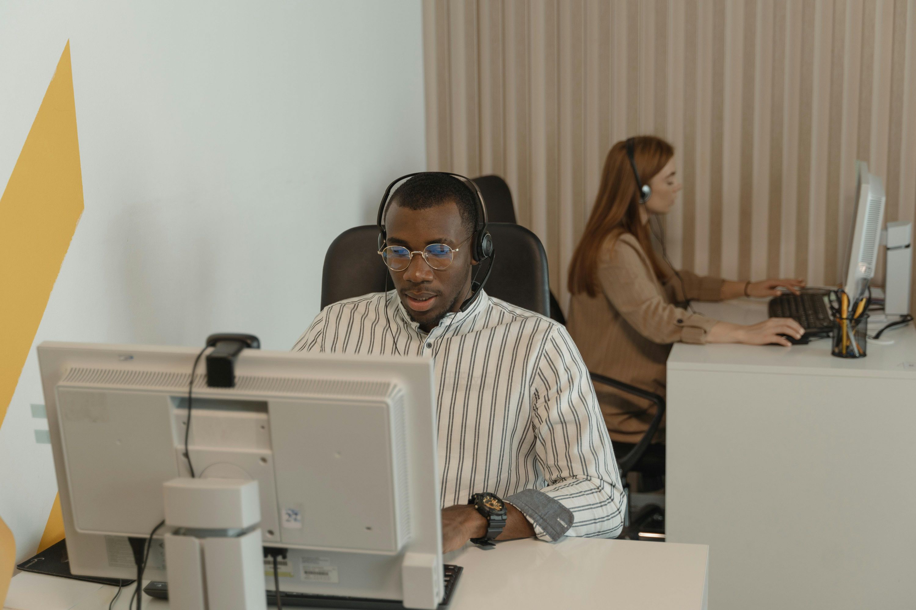 A concentrated young man in a striped shirt and glasses wearing a headset, working on a computer in an office, with a woman in the background.