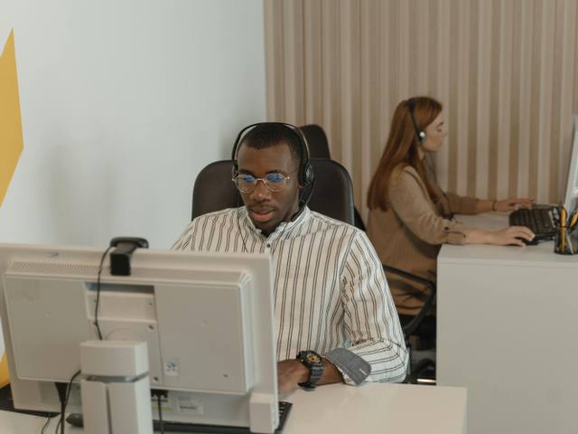 Remote Prompt Engineer at Work - Marketing Growth A concentrated young man in a striped shirt and glasses wearing a headset, working on a computer in an office, with a woman in the background.