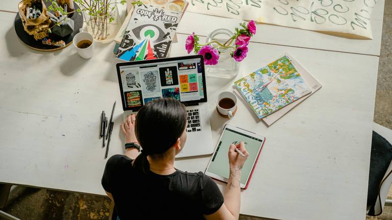 Creative Marketing Contractor for Outsourced Marketing Creative marketing contractor working on laptop and drawing tablet, surrounded by design tools and plants.