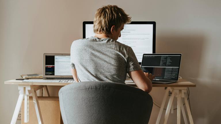 Outsourced Marketing Specialists Working Remotely Man working attentively at a desk with two laptops and a large monitor, demonstrating focus on digital tasks.