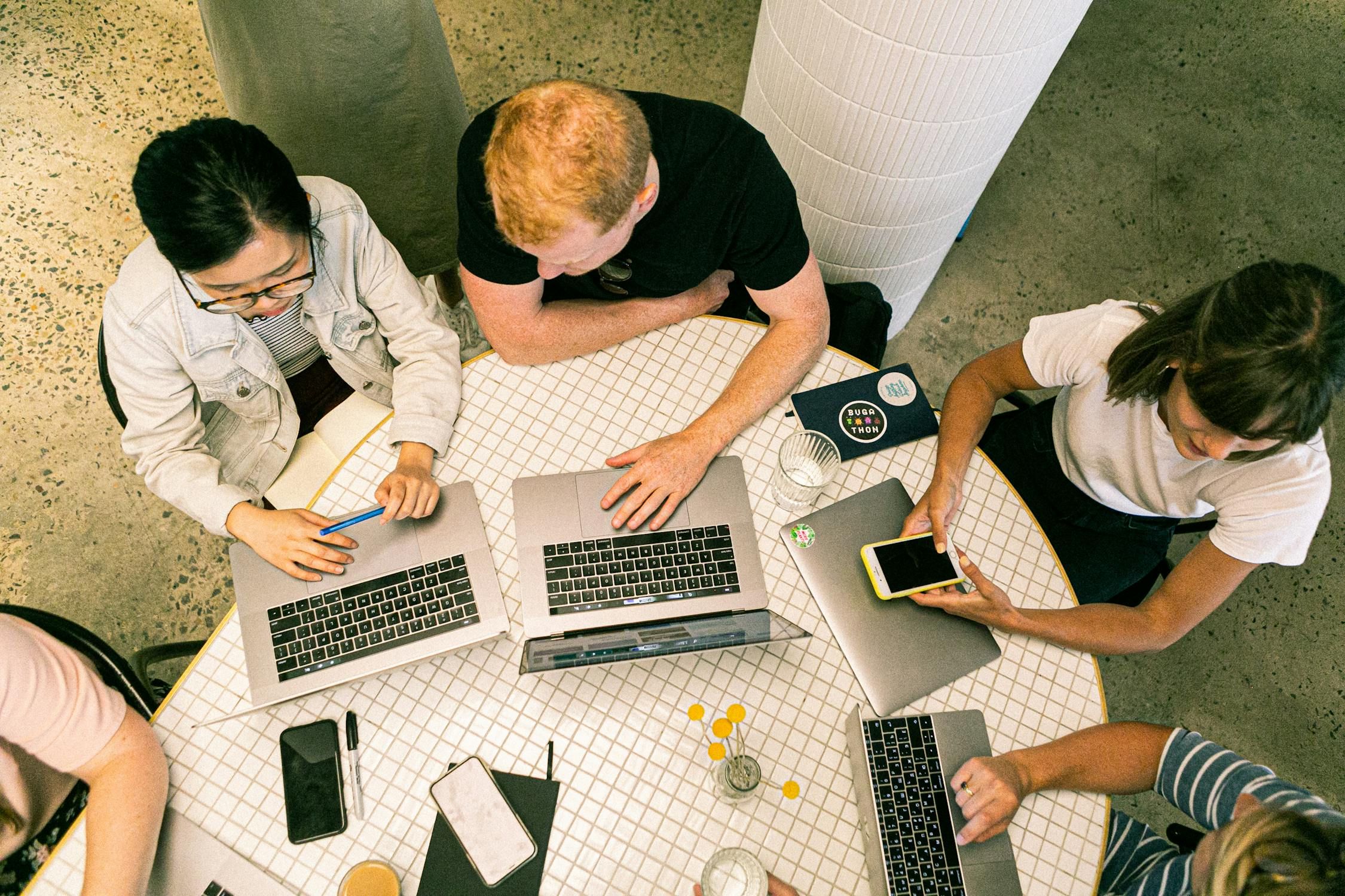 Overhead view of four marketing team members collaborating at a table with laptops, notebooks, and smartphones, actively working.