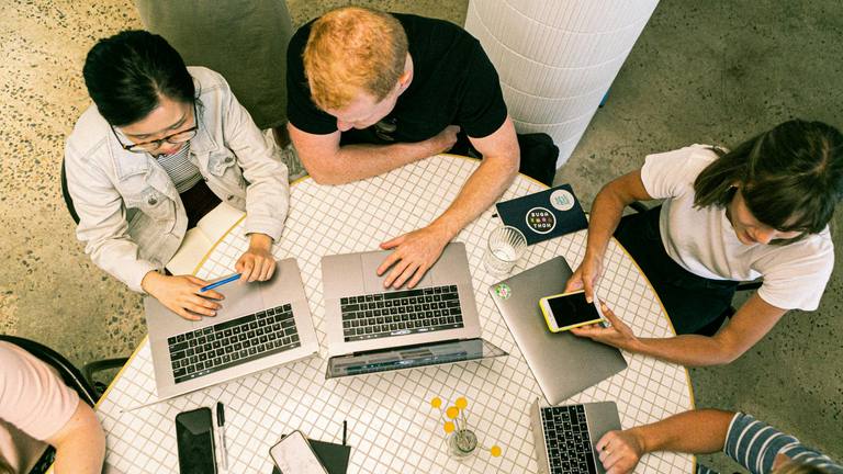 Marketing Team Collaboration - Roocruit Overhead view of four marketing team members collaborating at a table with laptops, notebooks, and smartphones, actively working.