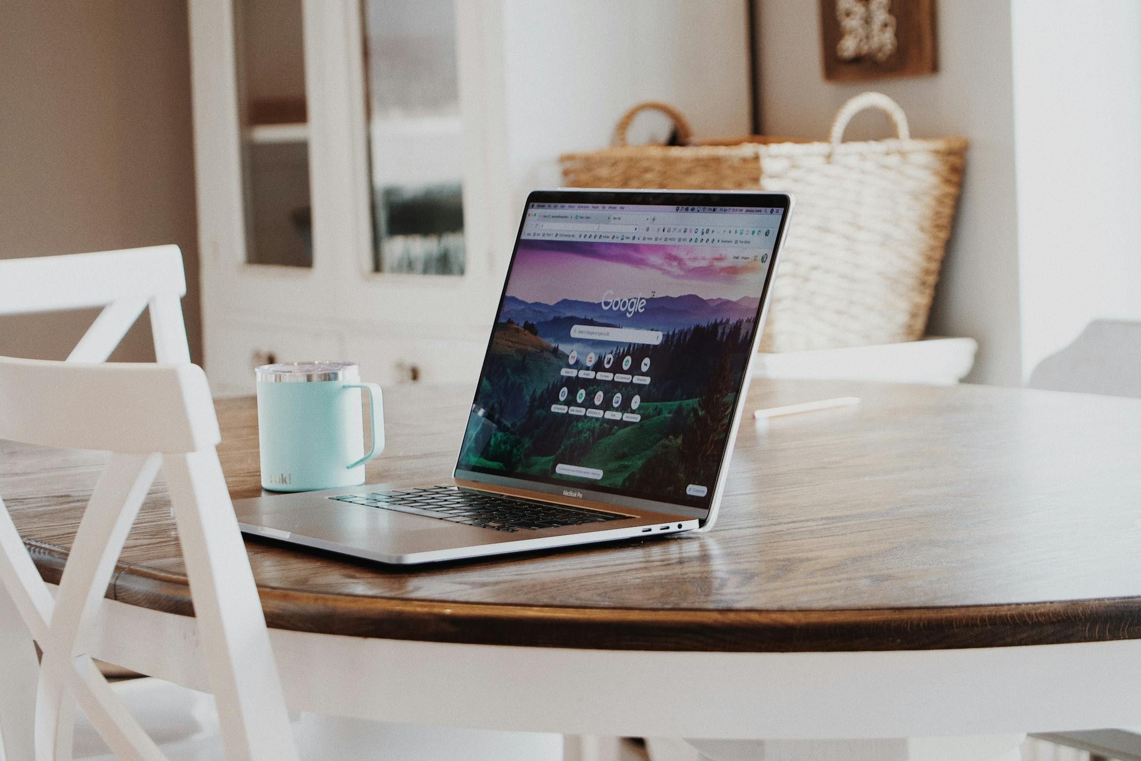 MacBook Pro laptop open to Google's homepage with a landscape background, accompanied by a teal travel mug on a wooden dining table, suggesting remote work.