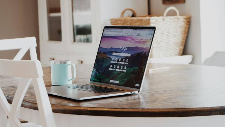 Google Ads Specialist - Remote Work Setup with MacBook Pro MacBook Pro laptop open to Google's homepage with a landscape background, accompanied by a teal travel mug on a wooden dining table, suggesting remote work.