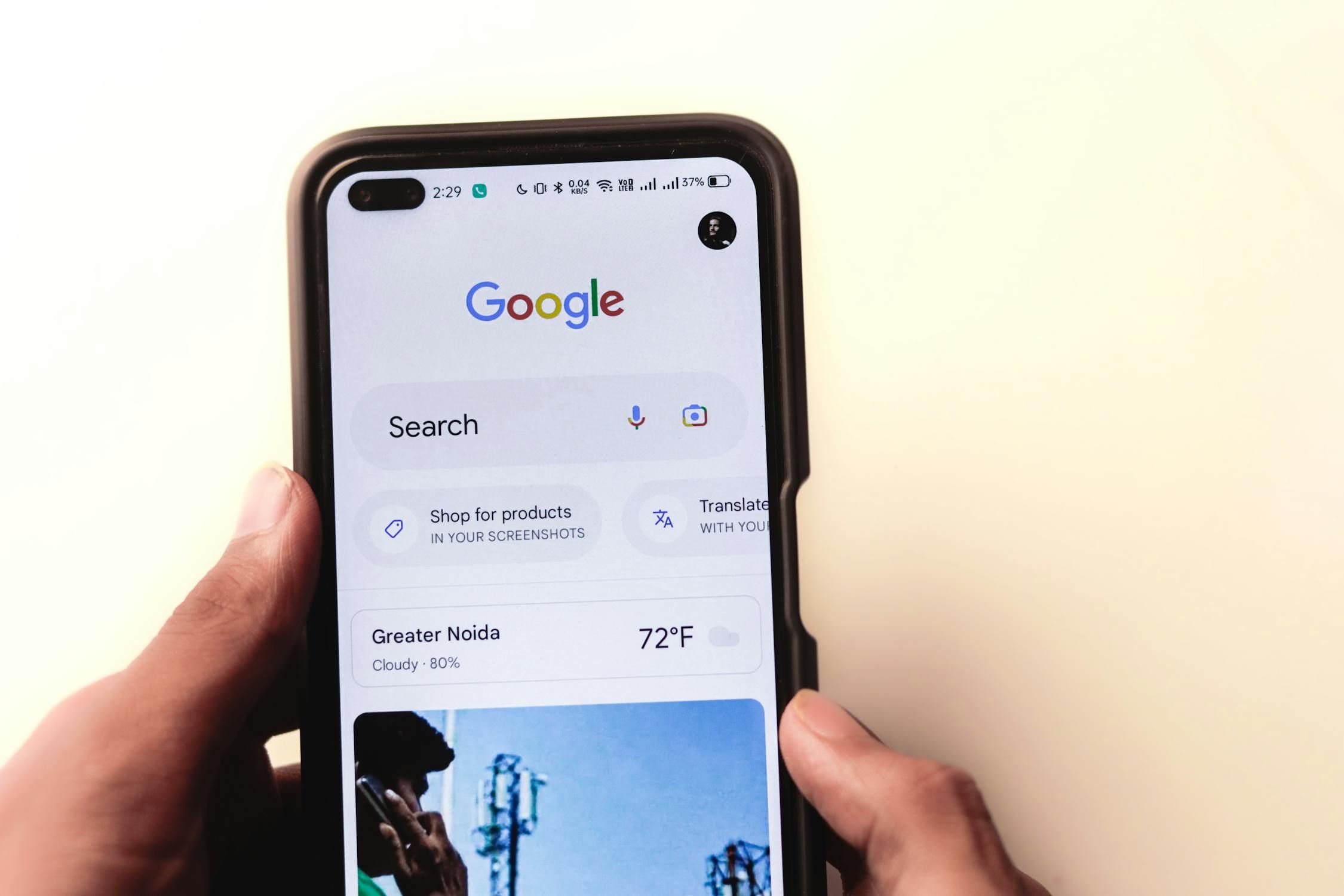 A hand holding a smartphone displaying the Google search page with local weather for Greater Noida and other features.