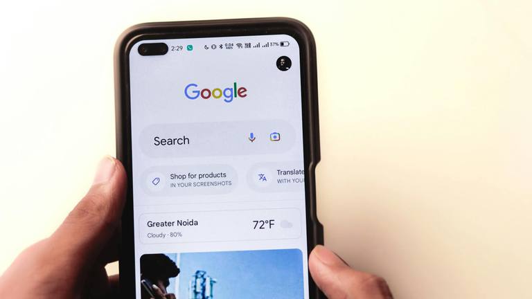 Google Search Page on Smartphone - Marketing Specialists A hand holding a smartphone displaying the Google search page with local weather for Greater Noida and other features.