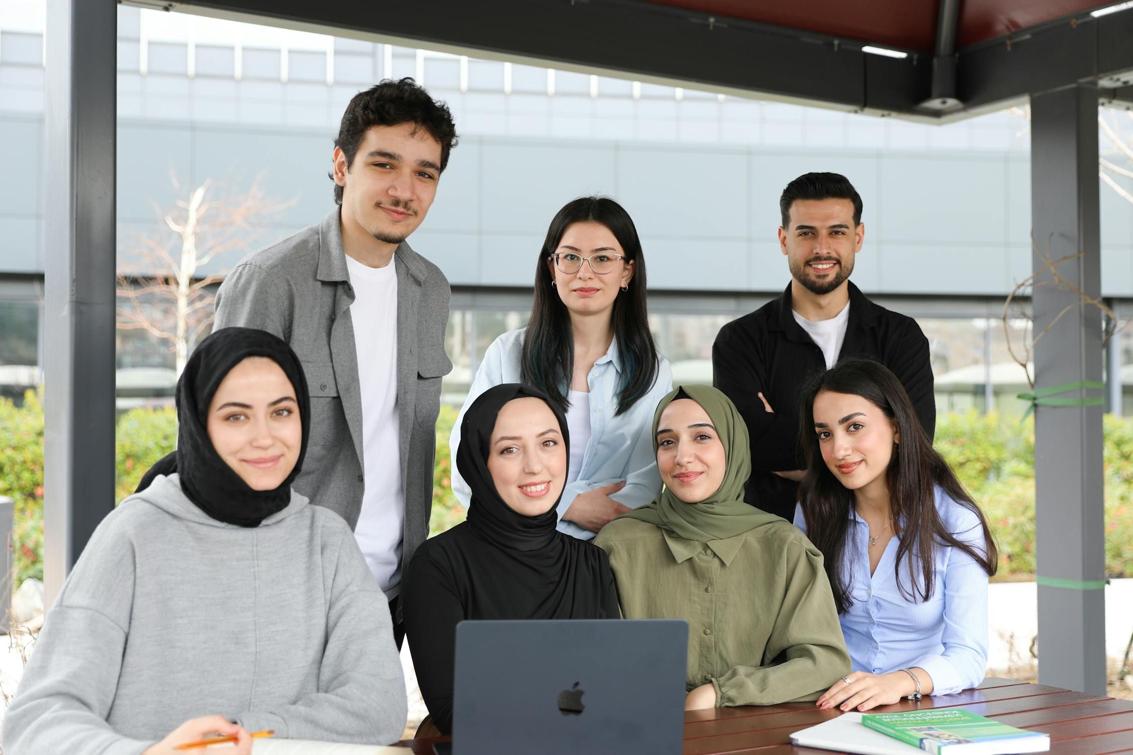 Diverse group of eight professional colleagues, including men and women with hijabs, smiling at the camera in a modern office setting.