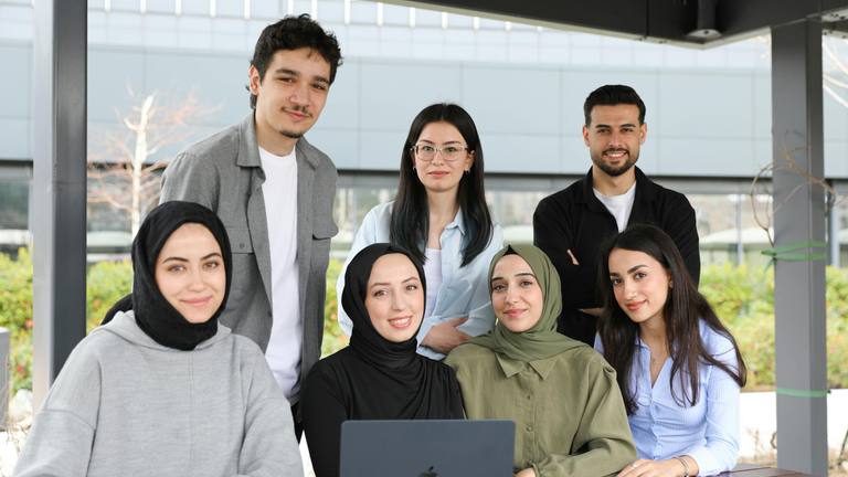 Remote Office Staff Team Collaboration | Marketing Diverse group of eight professional colleagues, including men and women with hijabs, smiling at the camera in a modern office setting.