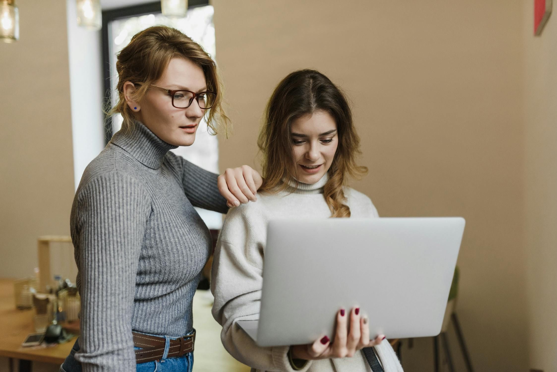 Two women, one in glasses, collaborating intently over a silver laptop in a brightly lit modern office setting.