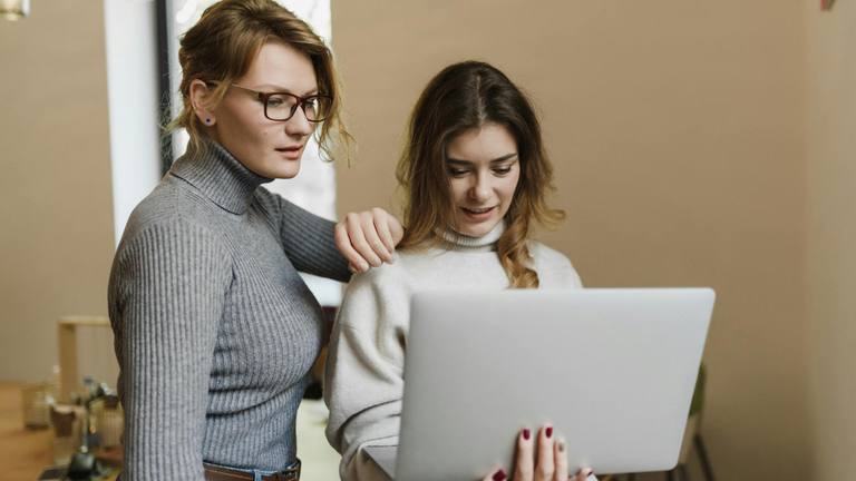SMEs Hiring Smarter: Collaborative Hiring Strategies for Small Businesses Two women, one in glasses, collaborating intently over a silver laptop in a brightly lit modern office setting.