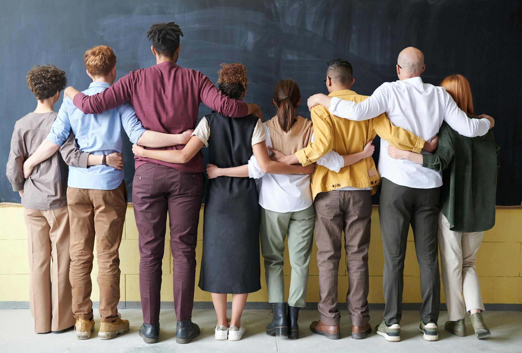 Diverse group of people standing arm-in-arm with their backs to the camera, looking at a chalkboard, symbolizing teamwork and unity.