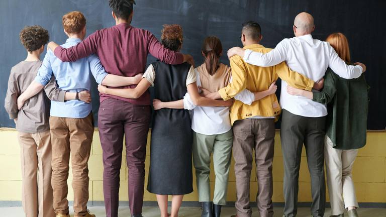 Outsourced Marketing Teamwork & Collaboration Diverse group of people standing arm-in-arm with their backs to the camera, looking at a chalkboard, symbolizing teamwork and unity.