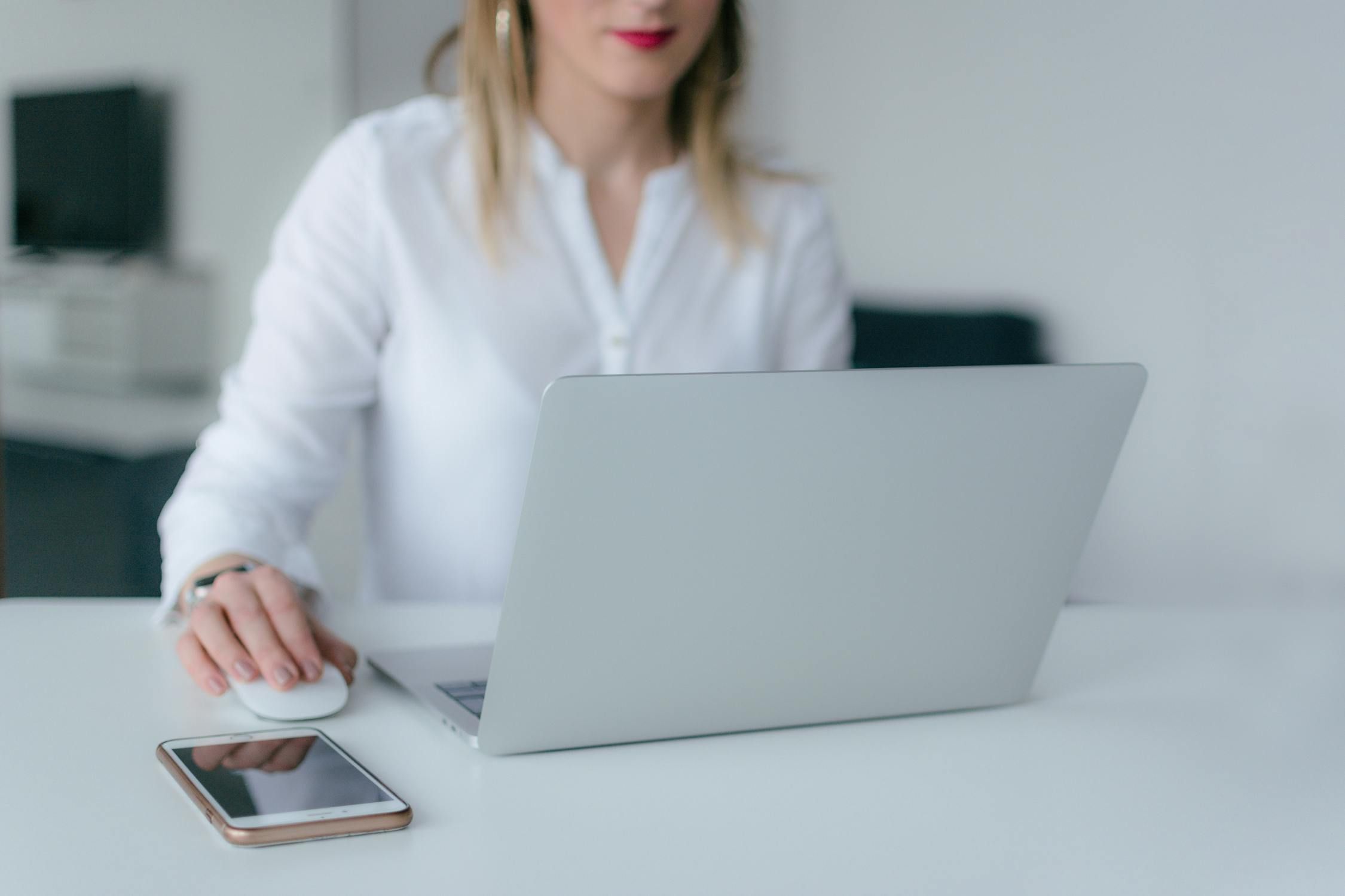 A woman working remotely at a desk, using a white mouse next to an open laptop and a smartphone.