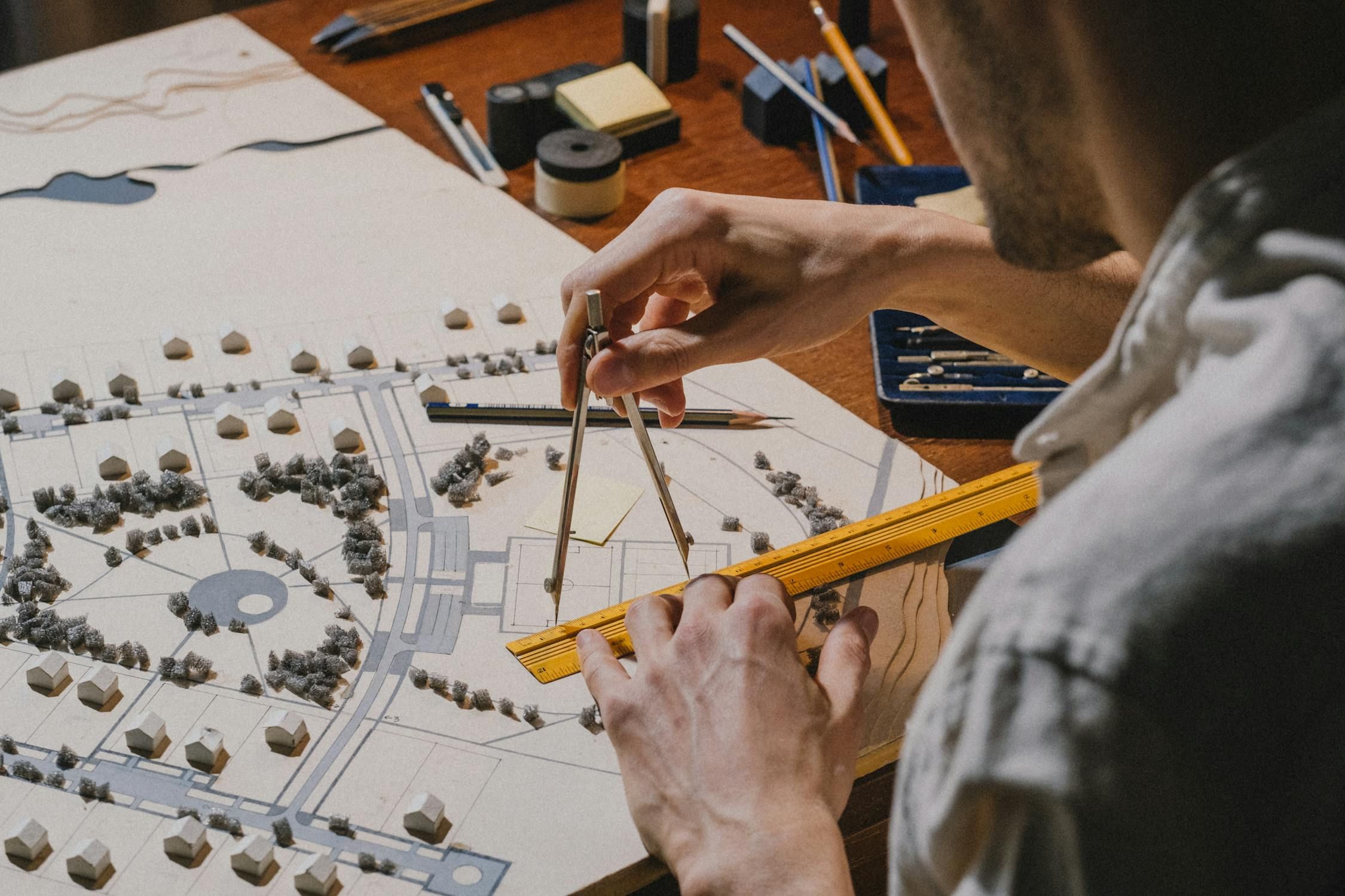 Architect using a compass and ruler to meticulously plan a model town with houses, trees, and roads.
