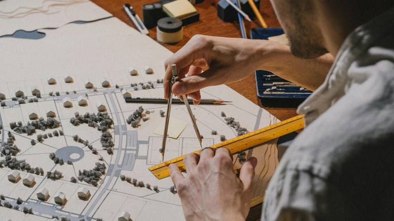 Strategic Marketing Planning & Freelance Marketers Architect using a compass and ruler to meticulously plan a model town with houses, trees, and roads.