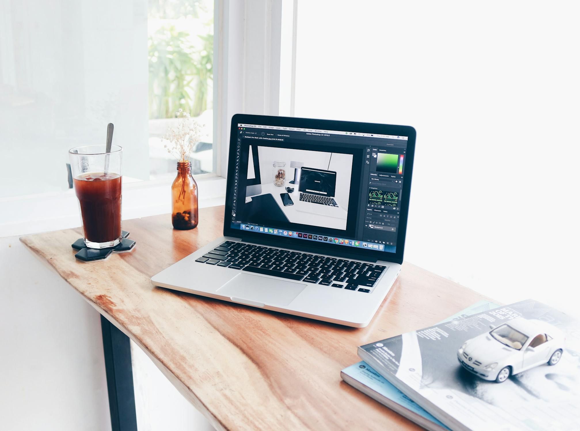 A MacBook Pro laptop open to Adobe Photoshop rests on a wooden desk next to iced coffee, a small vase, books, and a toy car.