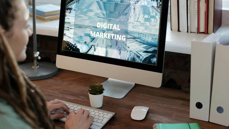 Digital Marketing Specialist Working Remotely A person with dreadlocks typing on a keyboard, looking at a computer monitor displaying the words "DIGITAL MARKETING" amidst a backdrop of abstract city buildings.