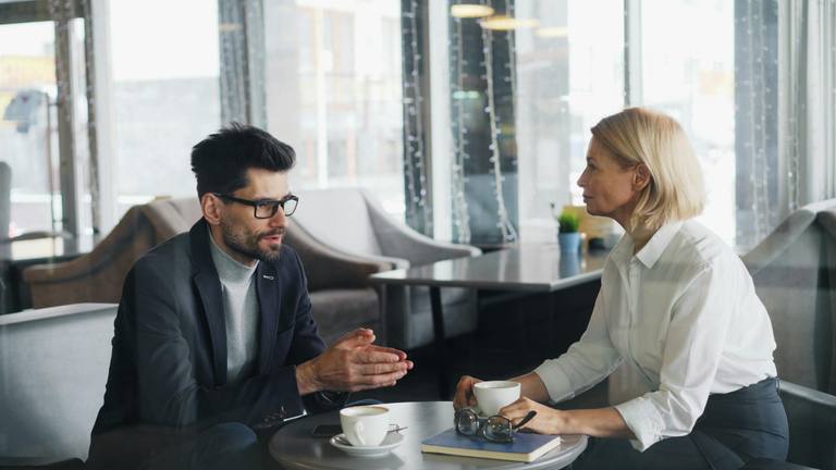 Business Colleagues Discussion | Flexible Remote Team Marketing Two colleagues, a man and a woman, engaged in a conversation at a table with coffee cups, suggesting a business meeting or discussion.