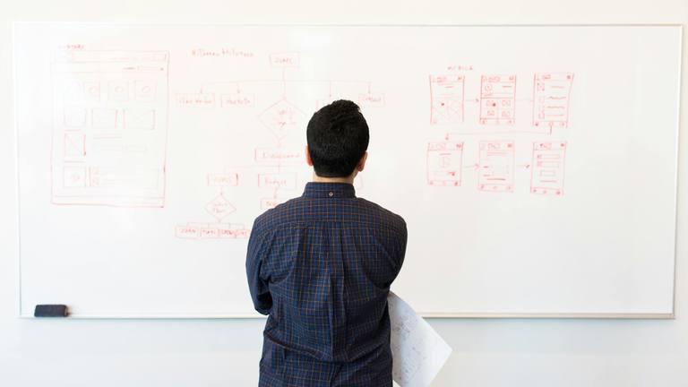 Marketing Strategy & Growth with Remote Talent A person viewed from behind, looking at a whiteboard filled with red marker diagrams, flowcharts, and mobile app wireframes.