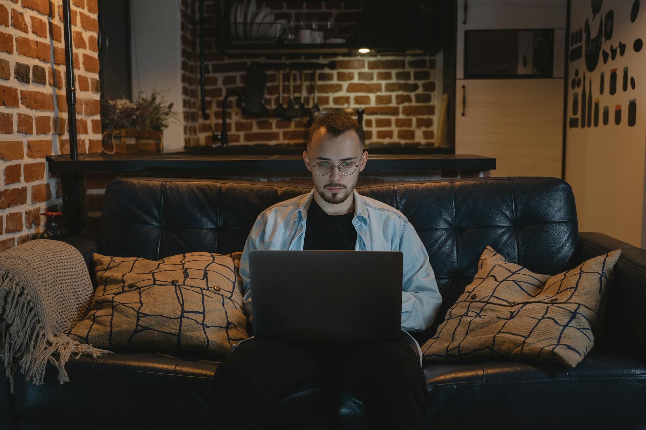 Young male professional with glasses working remotely on a laptop while relaxing on a dark leather couch in a modern loft.