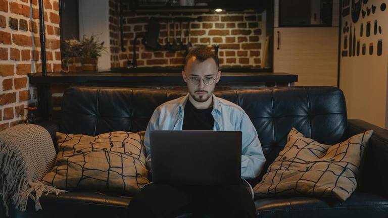 Remote Work Professional on Laptop for Marketing Young male professional with glasses working remotely on a laptop while relaxing on a dark leather couch in a modern loft.