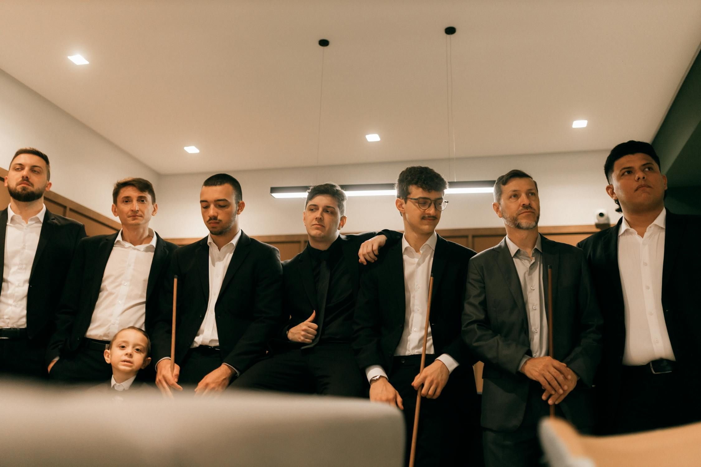 Seven men, one boy, all dressed in formal attire including suits and ties, stand or sit, looking upwards with serious expressions, holding pool cues.