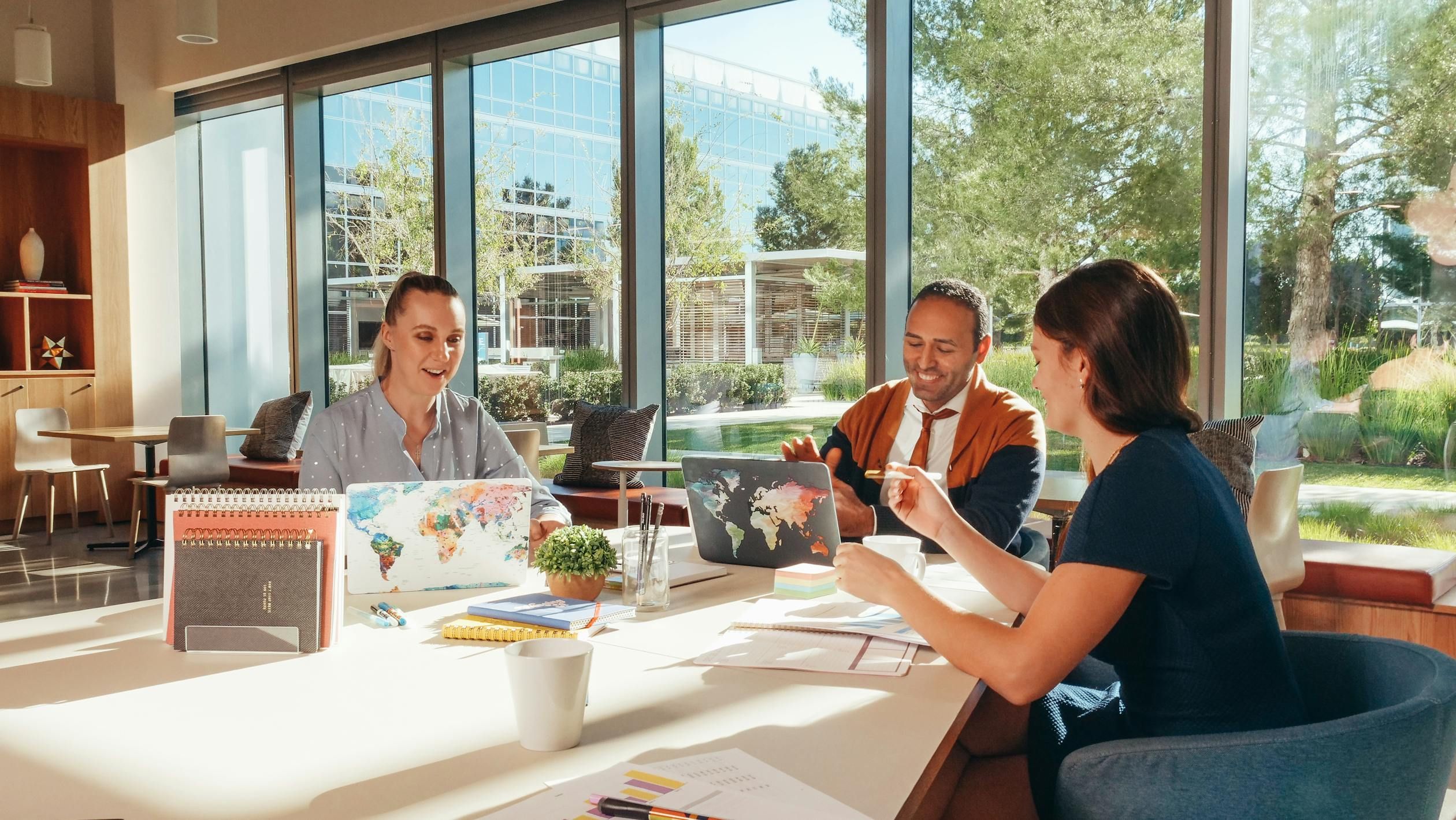 Three professionals collaborating around a bright desk with laptops featuring world map covers in a modern office with large windows overlooking trees.